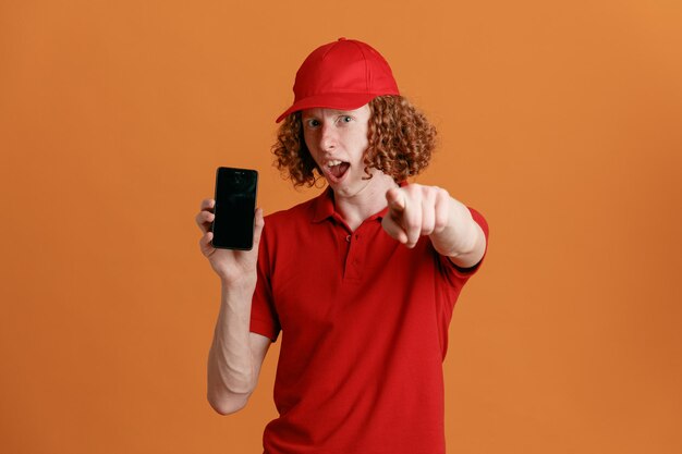 Lieferbote-Angestellter in roter T-Shirt-Uniform mit leerer Kappe, der das Smartphone zeigt, das mit dem Zeigefinger auf die Kamera zeigt, erstaunt und überrascht, wie er über orangefarbenem Hintergrund steht