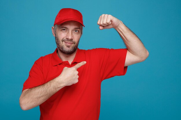 Lieferbote-Angestellter in roter T-Shirt-Uniform mit leerer Kappe, der mit selbstbewusstem Ausdruck in die Kamera blickt und die geballte Faust hebt, die auf seinen Bizeps zeigt, der über blauem Hintergrund steht