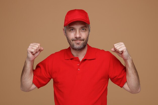 Lieferbote-Angestellter in roter T-Shirt-Uniform mit leerer Kappe, der mit selbstbewusstem Ausdruck in die Kamera schaut und die geballten Fäuste hebt, die über braunem Hintergrund stehen