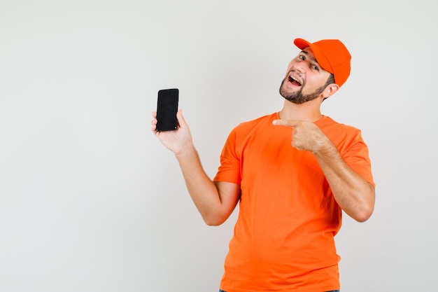 Lieferbote, der auf Handy in orangefarbenem T-Shirt, Mütze zeigt und fröhlich aussieht. Vorderansicht.