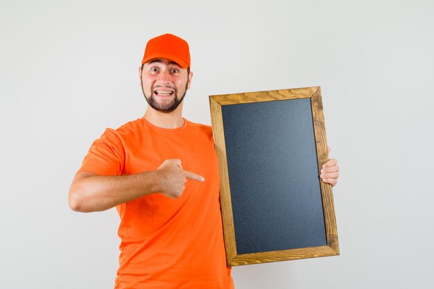 Lieferbote, der auf leere Tafel in orangefarbenem T-Shirt, Mütze zeigt und lustig aussieht. Vorderansicht.