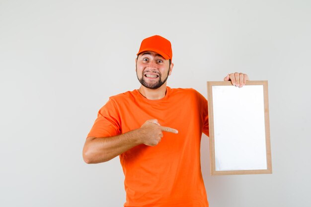Lieferbote, der auf leeren Rahmen in orangefarbenem T-Shirt, Mütze zeigt und lustig aussieht. Vorderansicht.