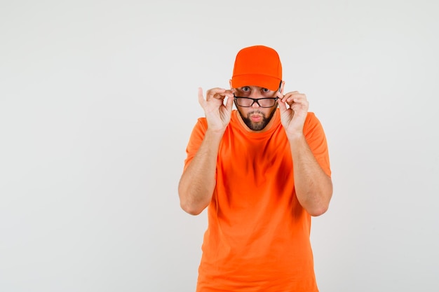 Lieferbote, der aufmerksam über die Brille in orangefarbenem T-Shirt, Mütze und überrascht schaut. Vorderansicht.