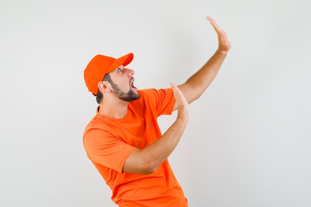 Lieferbote, der die Hände hebt, um sich in orangefarbenem T-Shirt, Mütze und verängstigtem Blick zu verteidigen, Vorderansicht.