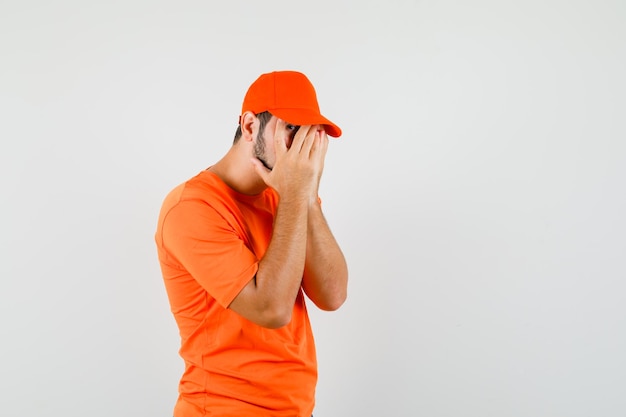 Lieferbote, der durch die Finger in orangefarbenem T-Shirt, Mütze schaut und verängstigt aussieht. Vorderansicht.