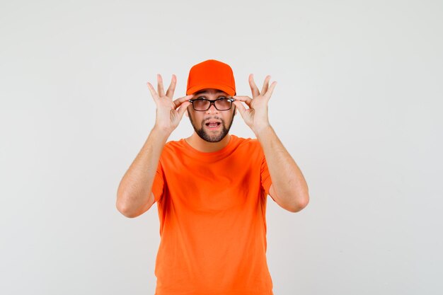 Lieferbote, der eine Brille in orangefarbenem T-Shirt, Mütze abnimmt und erstaunt aussieht, Vorderansicht.