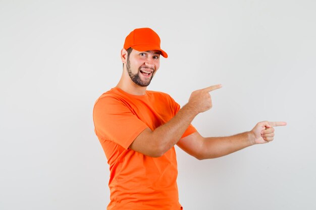 Lieferbote, der in orangefarbenem T-Shirt, Mütze und fröhlicher Miene auf die Seite zeigt. Vorderansicht.