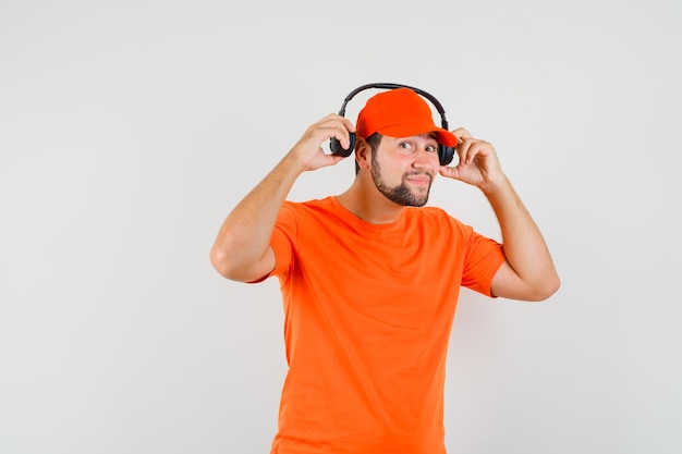 Lieferbote, der Kopfhörer in orangefarbenem T-Shirt, Mütze und fröhlichem Blick auszieht, Vorderansicht.