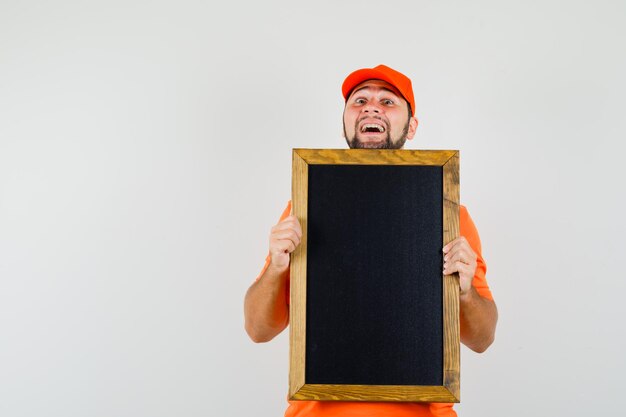 Lieferbote, der leere Tafel im orangefarbenen T-Shirt, in der Mütze hält und fröhlich aussieht. Vorderansicht.