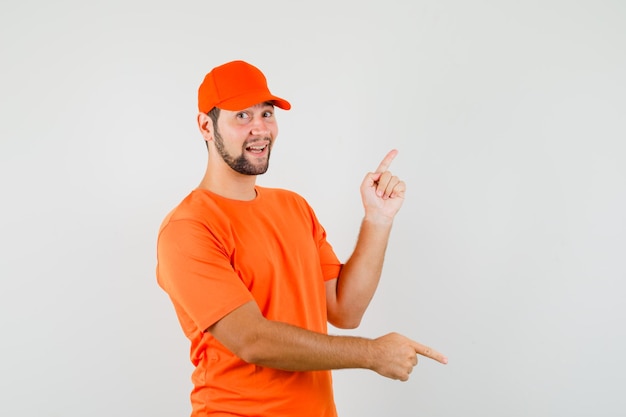 Lieferbote, der mit den Fingern in orangefarbenem T-Shirt, Mütze und fröhlichem Blick nach oben und unten zeigt. Vorderansicht.