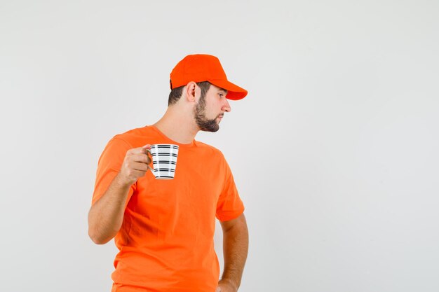 Lieferbote, der mit einer Tasse Getränk in orangefarbenem T-Shirt, Mütze und nachdenklichem Blick beiseite schaut, Vorderansicht.