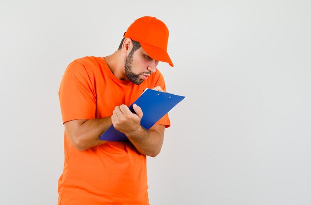 Lieferbote, der Notizen über die Zwischenablage in orangefarbenem T-Shirt, Mütze macht und beschäftigt aussieht, Vorderansicht.