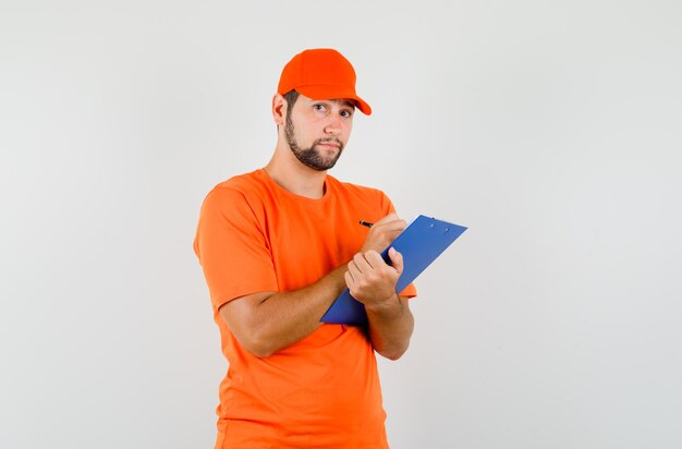 Lieferbote, der Notizen über die Zwischenablage in orangefarbenem T-Shirt, Mütze macht und vernünftig aussieht, Vorderansicht.