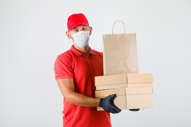 Lieferbote, der Pappkartons und Papiertüte in roter Uniform, medizinischer Maske, Handschuhen Vorderansicht hält.