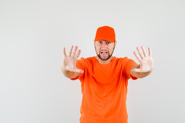 Lieferbote, der Stop-Geste in orangefarbenem T-Shirt, Mütze und verängstigtem Blick zeigt, Vorderansicht.