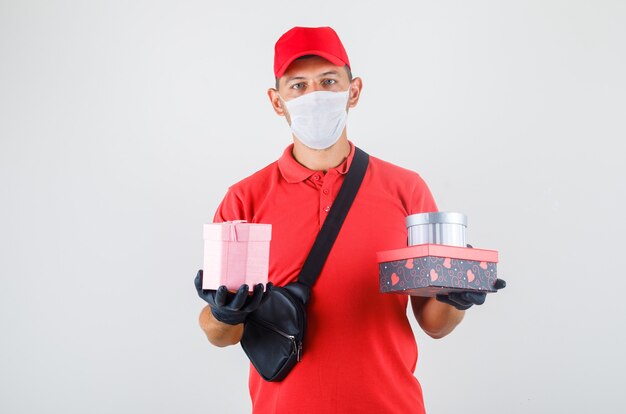 Lieferbote hält Geschenkboxen in roter Uniform, medizinischer Maske, Handschuhen Vorderansicht.