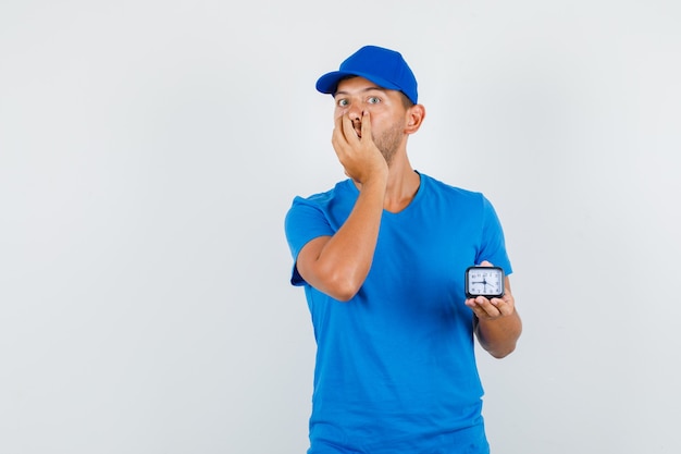 Lieferbote im blauen t-shirt, kappe, die uhr mit hand auf mund hält und aufgeregt schaut