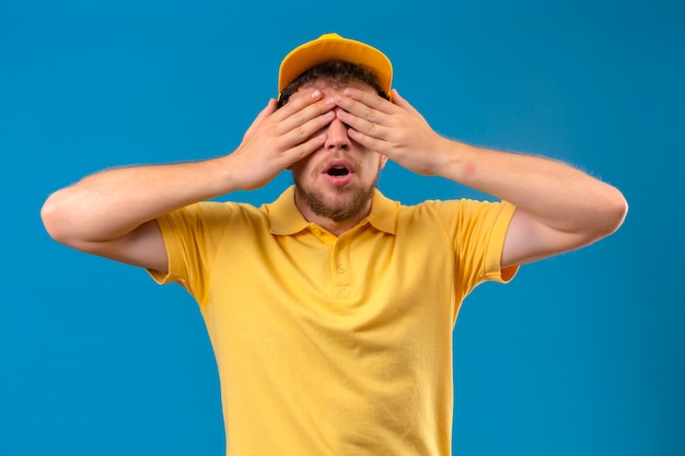 Lieferbote im gelben Poloshirt und in den Kappenkegelaugen mit den Händen für Überraschung, die auf isoliertem Blau stehen