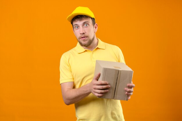 Lieferbote im gelben Poloshirt und in der Kappe, die Boxpaket hält, überrascht mit den Augen weit offen ope stehend auf isoliertem Orange