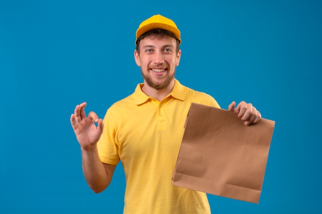 Lieferbote im gelben Poloshirt und in der Kappe, die Papierpaket halten, das freundlich tut, das ok Zeichen steht, das auf Blau steht