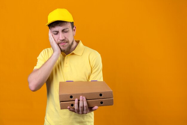 Lieferbote im gelben Poloshirt und in der Kappe, die Pizzaschachteln halten, die schlecht stehen mit Hand auf Wange stehen, die unter Zahnschmerzen auf Orange leiden