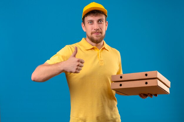 Lieferbote im gelben Poloshirt und in der Kappe, die Pizzaschachteln selbstzufrieden und glücklich hält, Daumen hoch stehend auf Blau zeigend