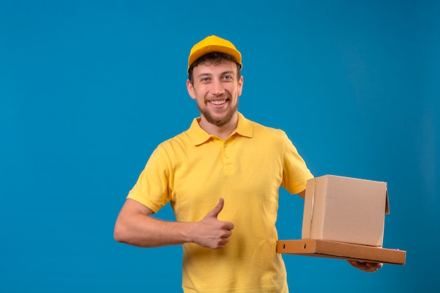 Lieferbote im gelben Poloshirt und in der Kappe, die Pizzaschachteln und Kastenverpackung selbstzufrieden halten und glücklich zeigen, Daumen hoch stehend auf Blau
