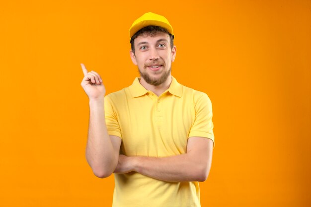 Lieferbote im gelben Poloshirt und in der Kappe lächelnd glücklich mit der Hand und dem Finger zur Seite zeigend auf lokalisiertem Orange stehend