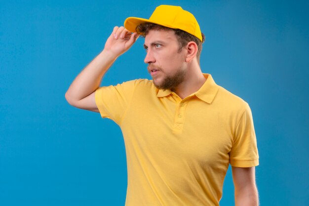 Lieferbote im gelben Poloshirt und in der Mütze, die beiseite schauen und Grußgeste machen, die seine Mütze auf isoliertem Blau berührt