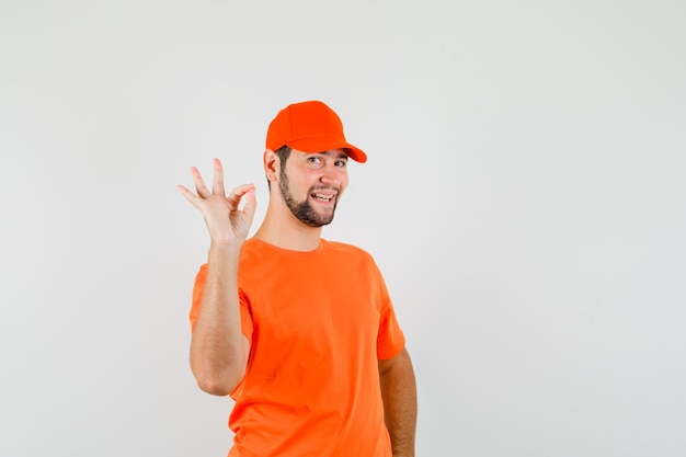 Lieferbote im orangefarbenen T-Shirt, Mütze, die eine ok Geste zeigt und fröhlich aussieht, Vorderansicht.