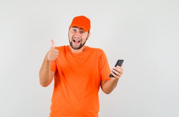 Lieferbote im orangefarbenen t-shirt, mütze, die handy mit daumen hoch hält und fröhlich aussieht, vorderansicht.