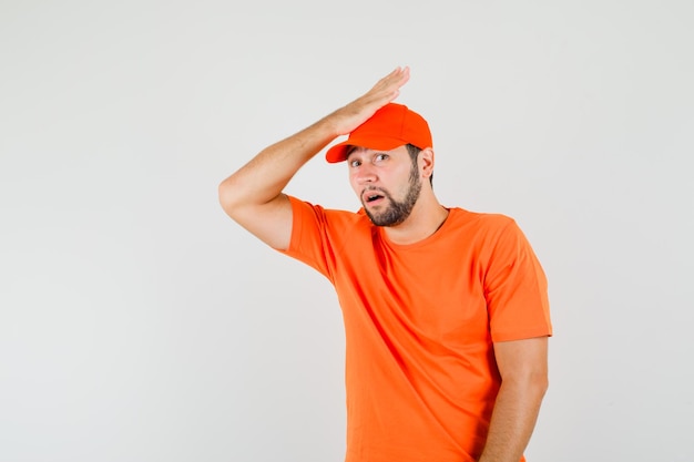 Lieferbote im orangefarbenen T-Shirt, Mütze mit Handfläche auf dem Kopf und auf der Suche nach Entschuldigung, Vorderansicht.