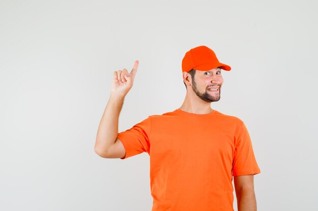 Lieferbote im orangefarbenen T-Shirt, Mütze zeigt mit dem Finger nach oben und sieht fröhlich aus, Vorderansicht.