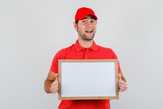 Lieferbote im roten T-Shirt, Kappe, die weiße Tafel hält und fröhlich schaut
