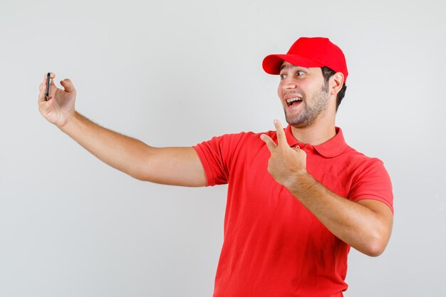 Lieferbote im roten T-Shirt, Mütze, die selfie mit V-Zeichen nimmt und fröhlich schaut