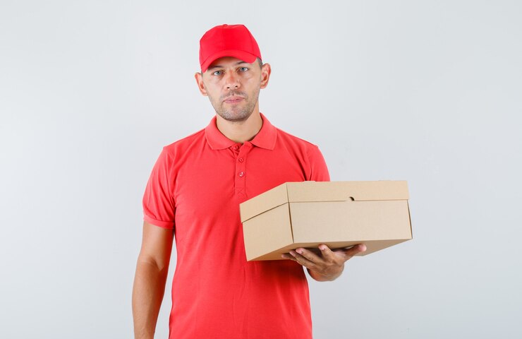 Kostenlose Foto lieferbote in der roten kappe und im t-shirt, die pappkarton halten und zuversichtlich schauen