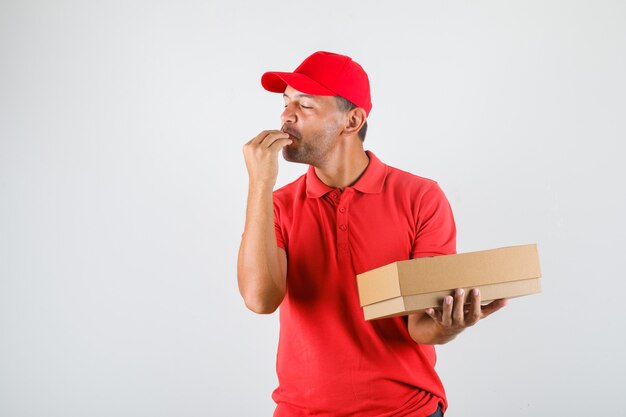 Lieferbote in der roten Uniform, die leckere Geste beim Halten der Pizzaschachtel macht