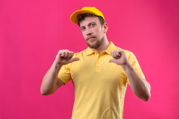 Lieferbote in gelbem Poloshirt und Mütze zeigen auf sich selbst, wobei beide Daumen selbstbewusst und zufrieden auf isoliertem Rosa stehen