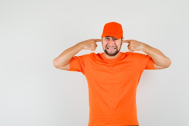 Lieferbote in orangefarbenem T-Shirt, Mütze, die Ohren mit den Fingern verstopft und genervt aussieht, Vorderansicht.