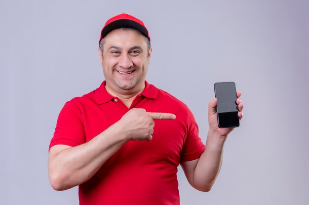 Lieferbote in roter Uniform und Kappe, die sein Smartphone zeigt, das mit dem Zeigefinger darauf zeigt und fröhlich auf Weiß steht
