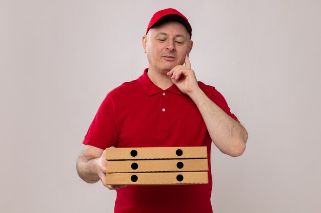 Lieferbote in roter Uniform und Mütze, die Pizzakartons hält und sie mit nachdenklichem Gesichtsausdruck ansieht und lächelt positiv denken