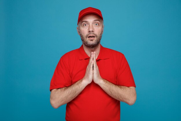 Lieferbote-Mitarbeiter in roter Kappe, leerem T-Shirt, Uniform, Blick in die Kamera, verwirrt, Handflächen zusammenhaltend, wie betend, über blauem Hintergrund stehend