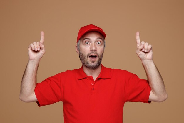Lieferbote-Mitarbeiter in roter Kappe, leerem T-Shirt, Uniform, die mit Zeigefingern nach oben zeigt und in die Kamera blickt, die überrascht über braunem Hintergrund steht