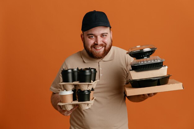 Lieferbote-mitarbeiter in schwarzer mütze und leerer t-shirt-uniform mit kaffeetassen und lebensmittelbehältern, die glücklich und positiv lächelnd freundlich über orangefarbenem hintergrund in die kamera blicken