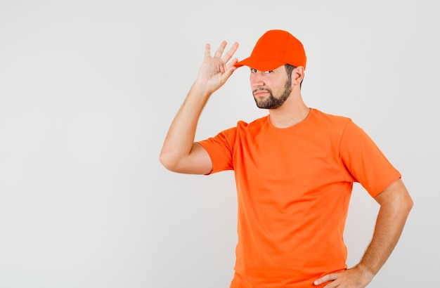 Lieferbote posiert mit den Fingern auf der Mütze im orangefarbenen T-Shirt und sieht selbstbewusst aus. Vorderansicht.