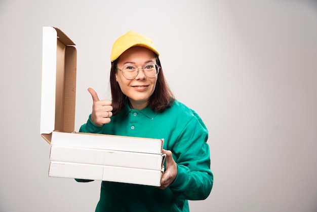 Lieferfrau, die einen Daumen zeigt und Pappkartons Pizza auf einem Weiß hält Foto in hoher Qualität