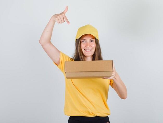 Lieferfrau, die Finger auf Pappkarton im gelben T-Shirt, in der Hose, in der Kappe zeigt und fröhlich schaut