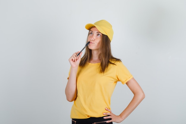 Lieferfrau, die Stift auf Lippen im gelben T-Shirt, in der Hose, in der Mütze hält und nachdenklich schaut.