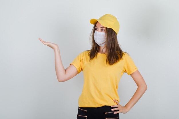Lieferfrau hält offene Handfläche in T-Shirt, Hose, Mütze und Maske