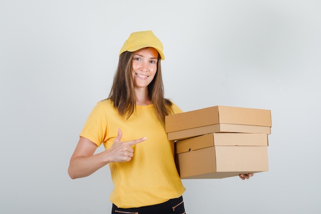 Lieferfrau im gelben T-Shirt, in der Hose, in der Kappe, die Finger auf Pappkartons zeigt und froh aussieht